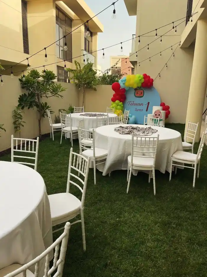 Round tables in white linen and chiavari chairs on a lawn strung with festoon lights, a first birthday backdrop and balloon garland behind them