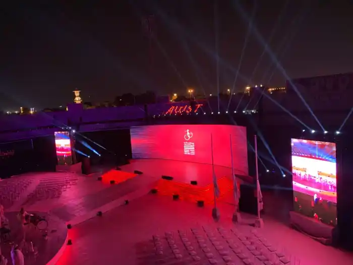 A curved stadium stage washed in red with beam lights fanning across the night sky