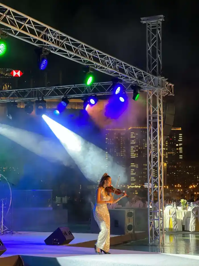 A violinist in a sequinned gown playing in a spotlight on an outdoor stage, city towers behind