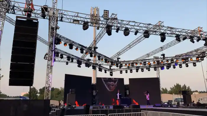 A large outdoor festival stage at dusk with line array speakers, truss lighting and performers on stage